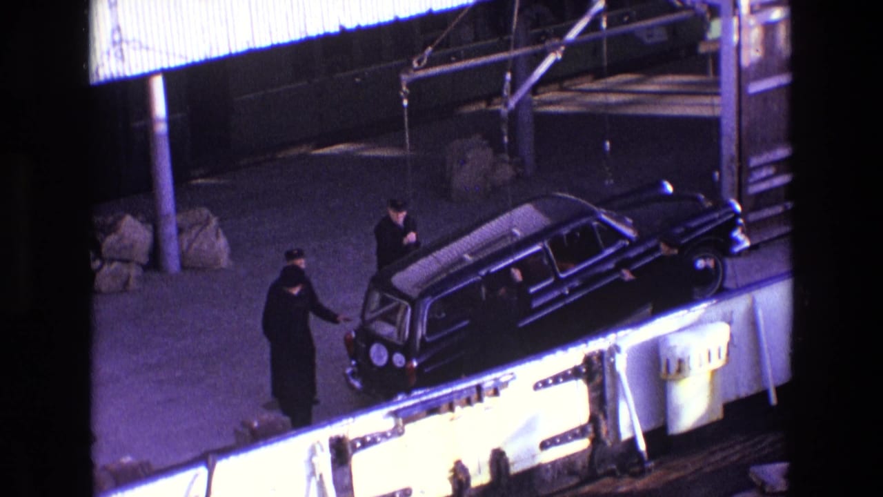 The black estate car is lowered beside an industrial platform while men in dark clothing guide it from the rear and side.