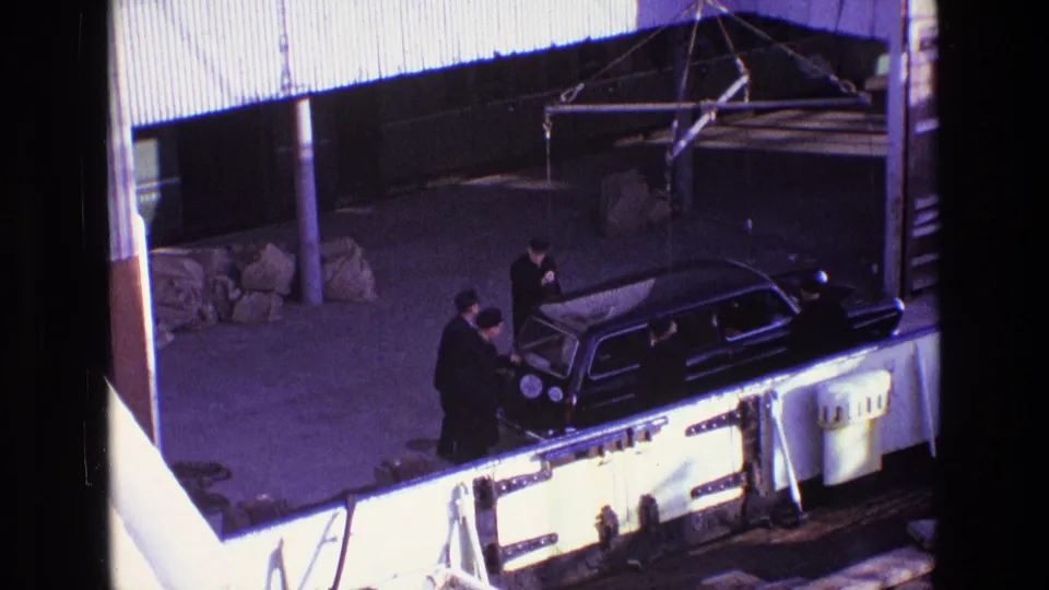 Men in dark coats cluster around the rear of the black estate car as it is positioned near a bright platform edge.
