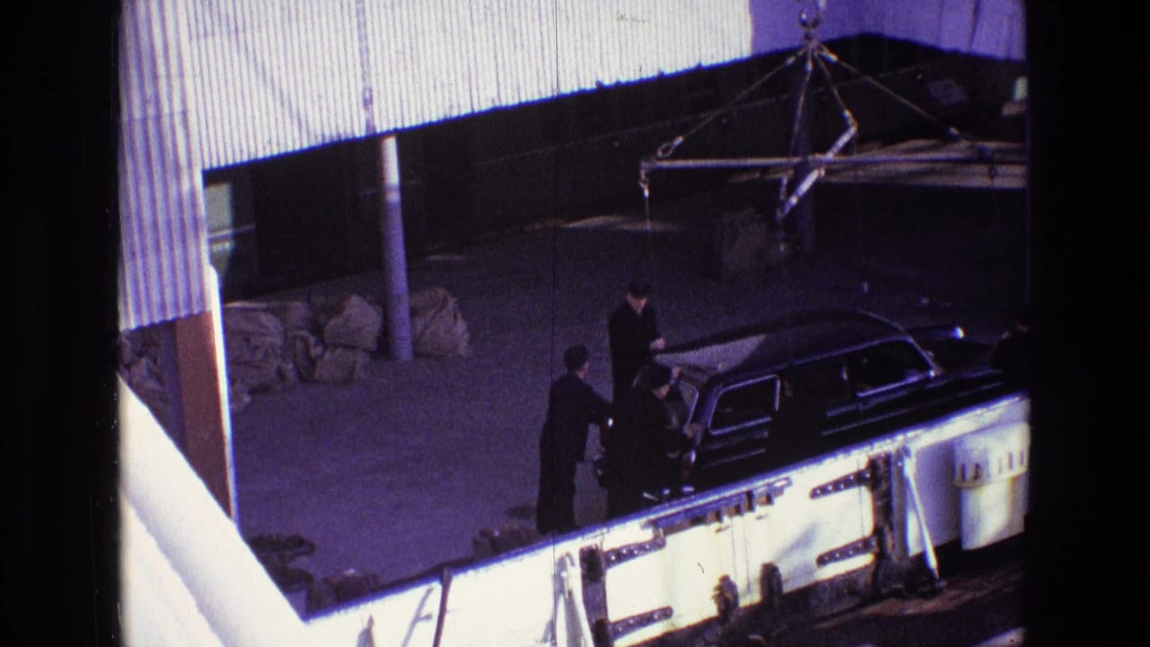 Black hearse seen from above with suited men standing beside it near a covered structure.