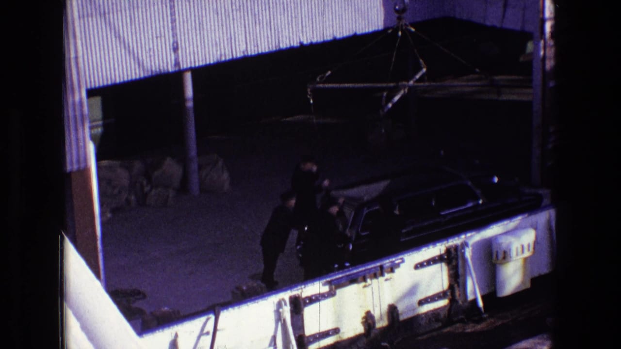 Men in dark suits stand beside a black hearse viewed from above near a building edge.