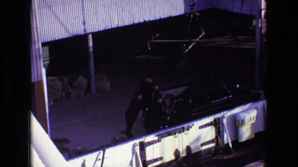 Elevated 1969 Ireland home-movie view of suited men beside a black hearse, with the scene partly framed by nearby structures.