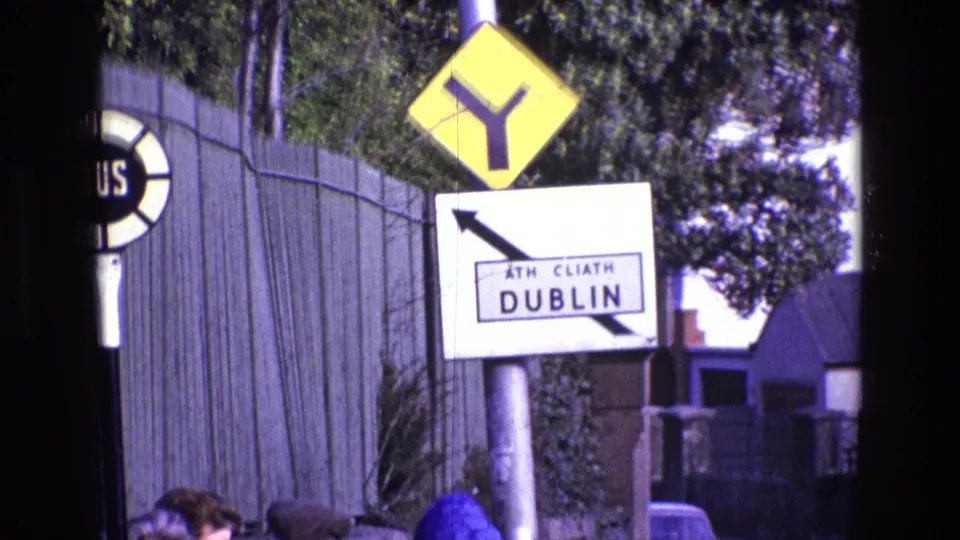 1969 Ireland roadside frame with a Dublin direction sign, yellow Y-junction sign, partial bus stop marker, fencing, trees, and street background.