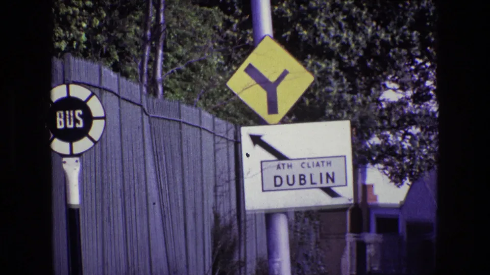1969 Ireland 8mm frame showing Dublin road signage, a yellow Y-junction sign, a bus stop marker, fence, trees, and a nearby building roofline.