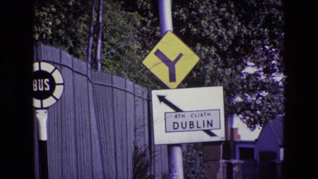 1969 Dublin roadside view with a circular BUS stop sign, ATH CLIATH DUBLIN direction sign, and yellow Y-intersection warning sign by trees and fencing.
