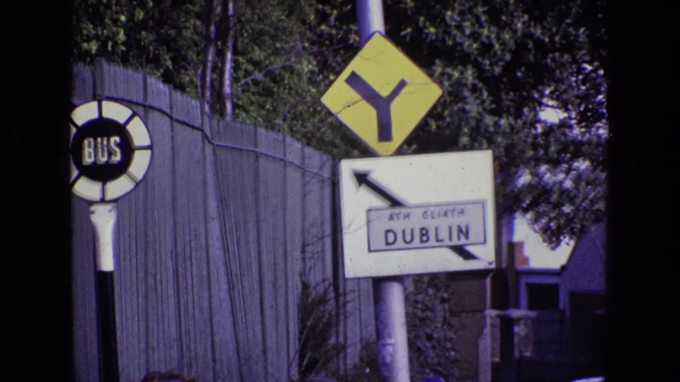 1969 Dublin roadside frame with the circular BUS sign, yellow Y-intersection marker, and ATH CLIATH DUBLIN direction sign seen through 8mm grain.