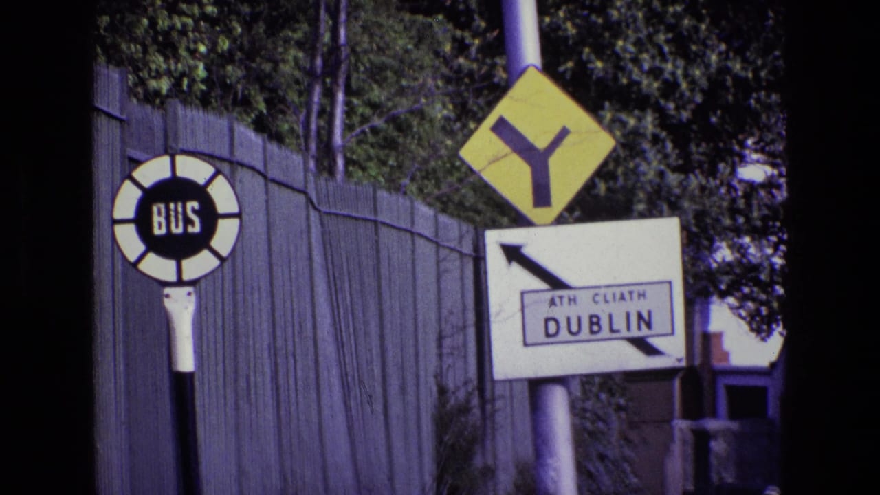 1969 Ireland 8mm frame showing a circular bus stop sign, yellow road warning sign, and Dublin direction sign near a fence.