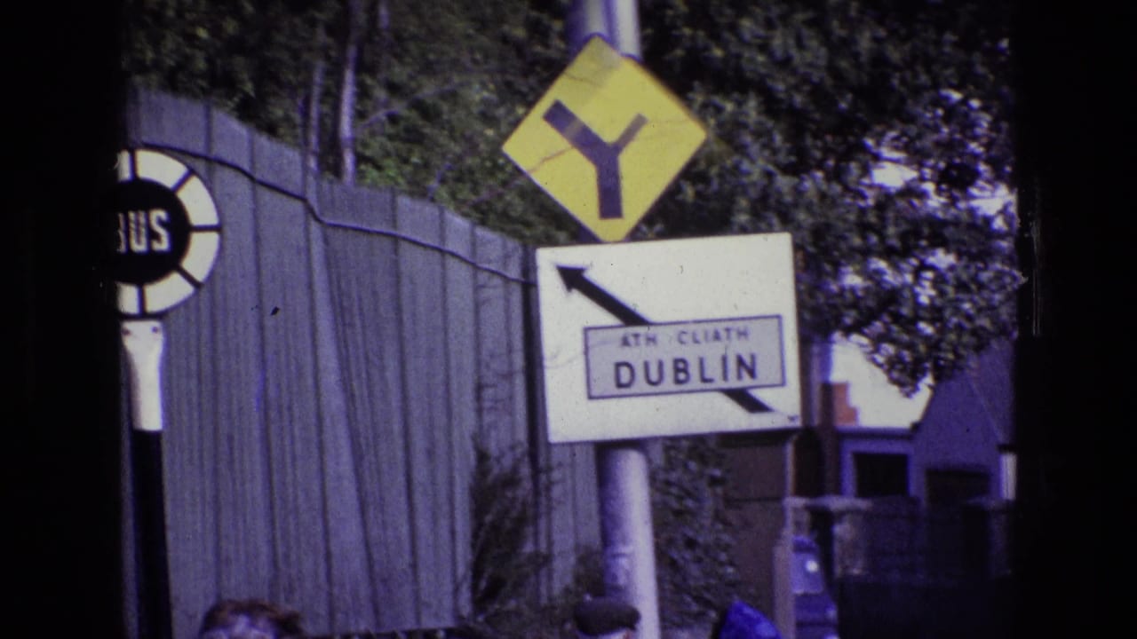 1969 Ireland roadside view with Dublin signage, a bus stop marker, warning sign, fence, and partially visible people at the bottom edge.