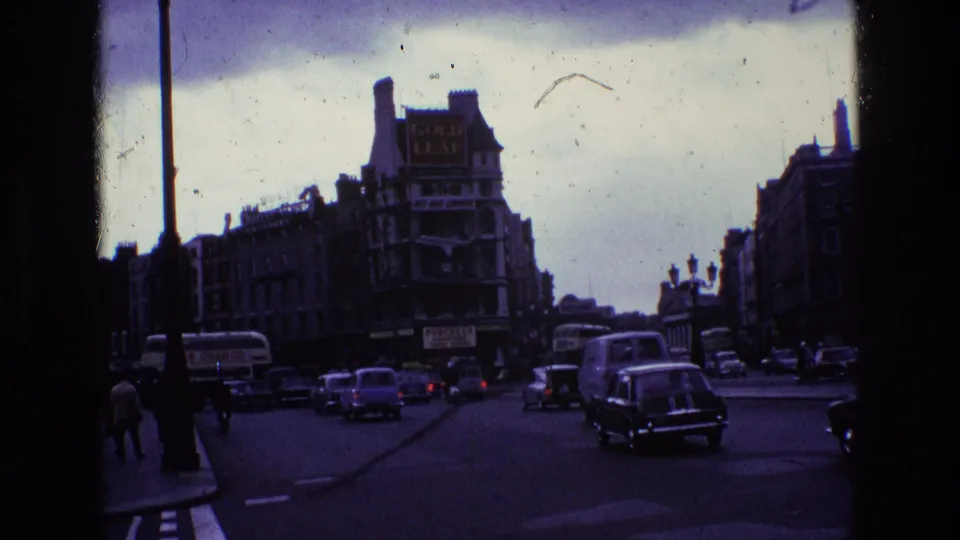 1969 Dublin street scene with period cars, pedestrians, shopfront signage, streetlights, and dark handheld 8mm exposure.