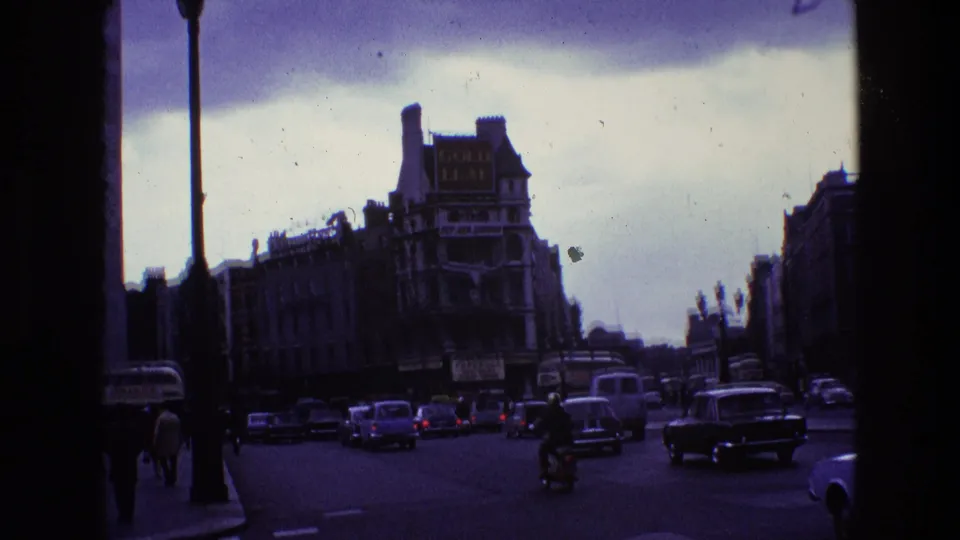 1969 Dublin traffic view showing classic cars, a motorcyclist, pedestrians, streetlights, and period commercial buildings.