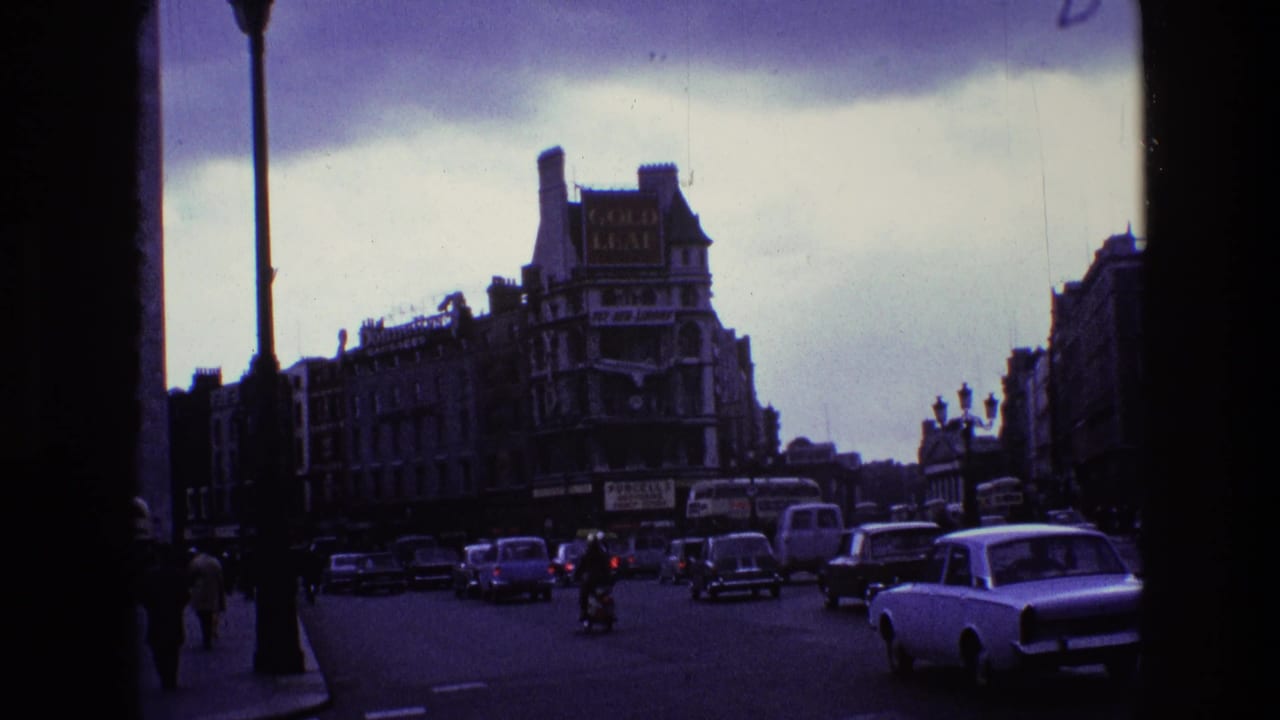 1969 Ireland street view framed by dark film edges, with vintage cars, pedestrians, tall lamps, and commercial signage.