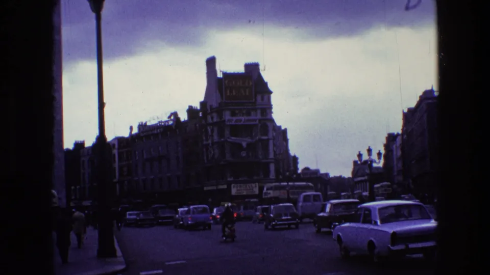 1969 Ireland street view framed by dark film edges, with vintage cars, pedestrians, tall lamps, and commercial signage.