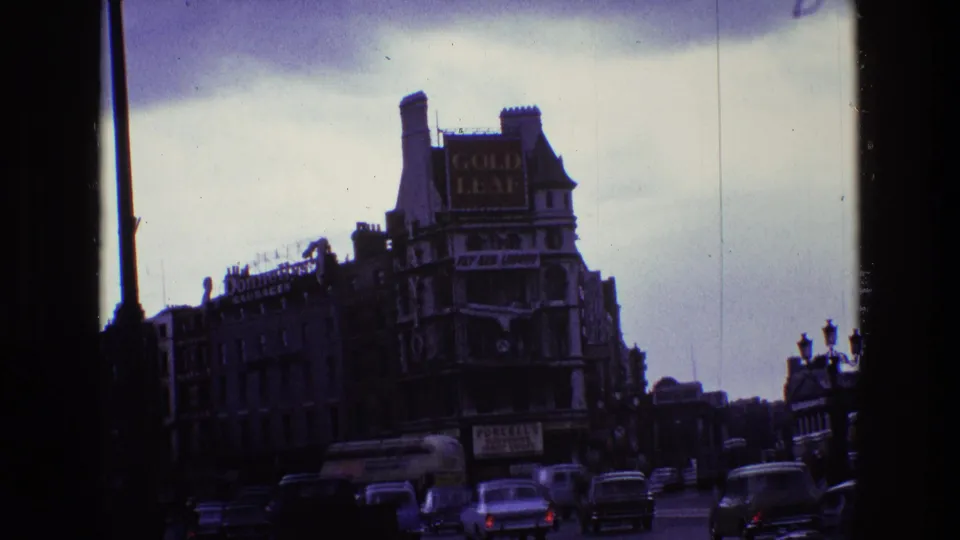 1969 Ireland urban traffic scene with vintage cars, streetlamps, pedestrians, and a large sign on a corner building.
