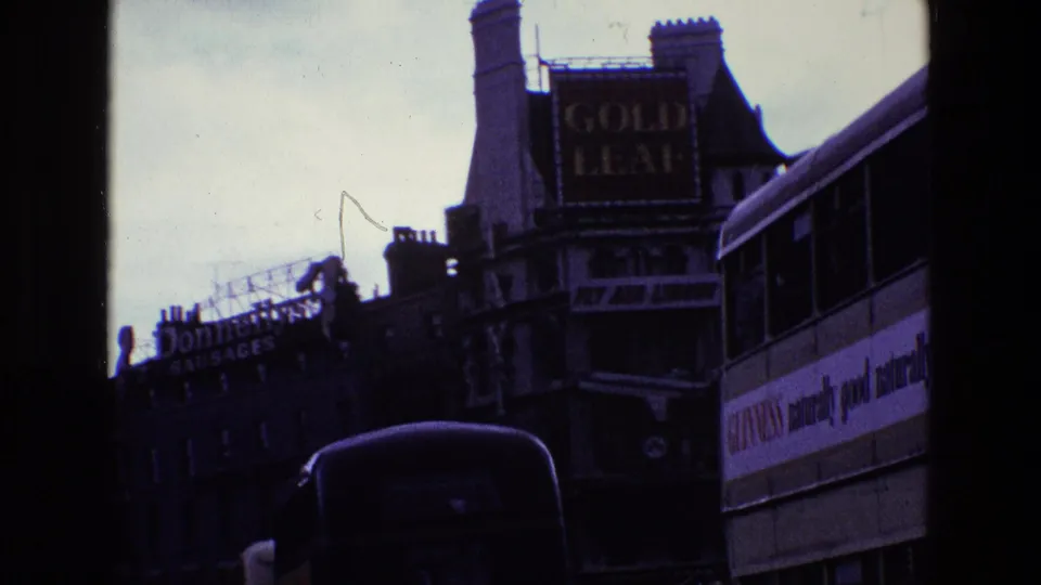 1969 Ireland city traffic scene with a white double-decker bus, dark car, Gold Leaf sign, and Donnelly's Sausages sign.