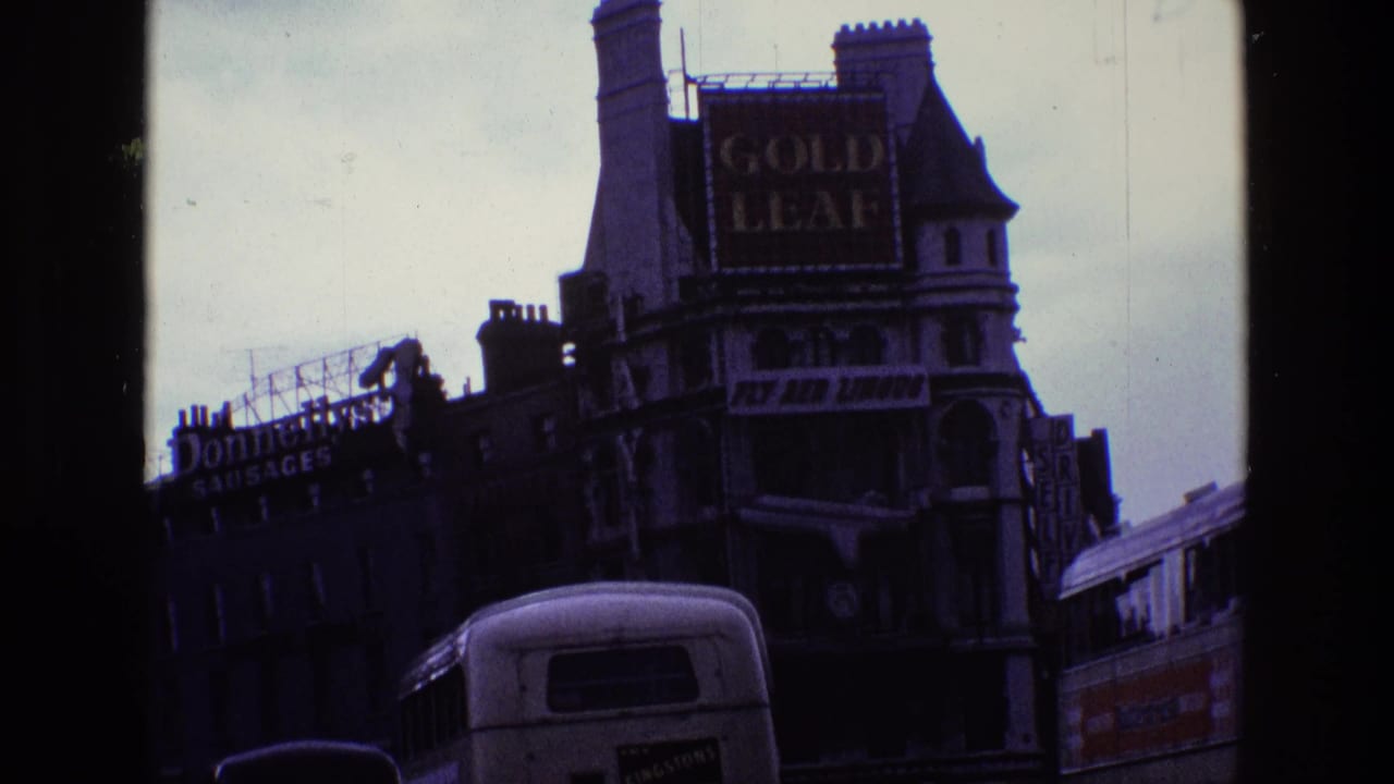 1969 Ireland home-movie frame showing a street corner facade with Gold Leaf and Fly Aer Lingus signs above passing buses.