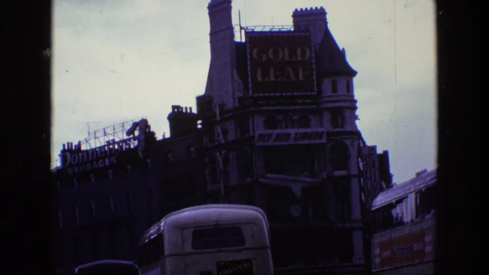 1969 Ireland home-movie frame showing a street corner facade with Gold Leaf and Fly Aer Lingus signs above passing buses.