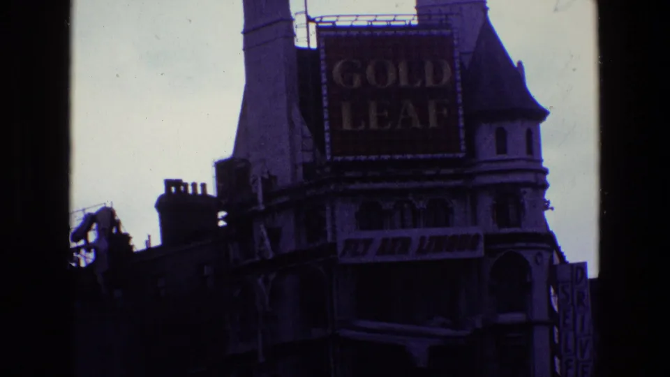 Closer 1969 Ireland frame centered on Gold Leaf and Fly Aer Lingus signs, with a darker street building and partial vertical sign.