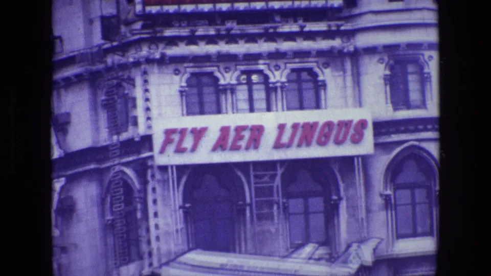1969 Ireland 8mm frame showing a building facade with a red FLY AER LINGUS sign and a model airplane below it.