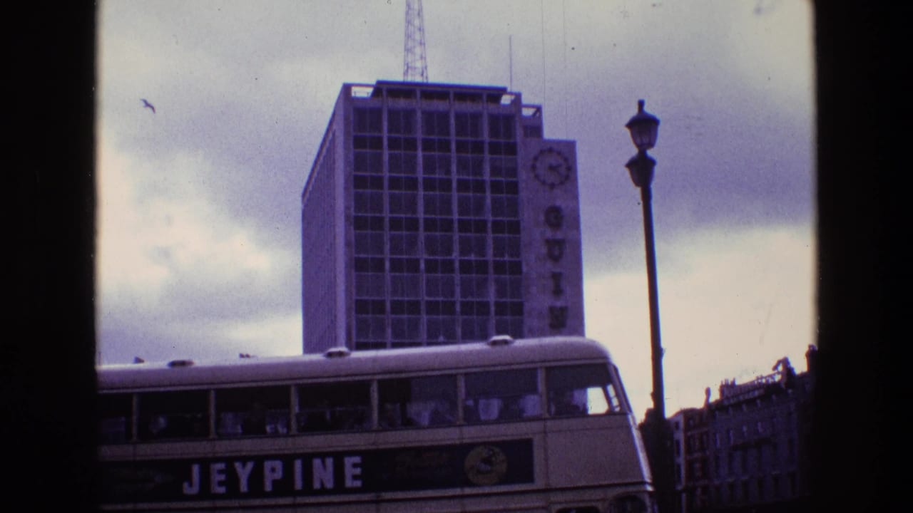 1969 8mm frame of a Jeypine double-decker bus moving past a tall Guinness building with a clock sign and street lamp.