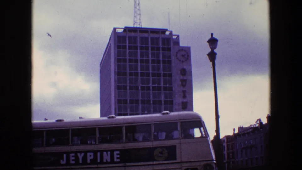 1969 8mm frame of a Jeypine double-decker bus moving past a tall Guinness building with a clock sign and street lamp.