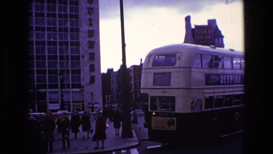 Street-level 8mm view of pedestrians near the Guinness building while a Jeypine double-decker bus turns beside the sidewalk.