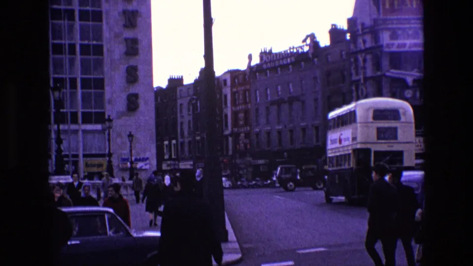 1969 Ireland street view with pedestrians near a crossing, shopfronts, cars, and a double-decker bus seen from a moving vehicle.