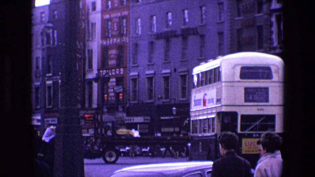 Double-decker bus passes storefronts as pedestrians stand near a busy street corner.