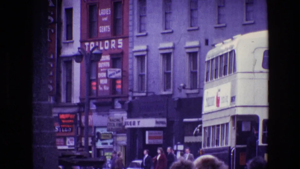 1969 Ireland street-level frame with storefront lettering, pedestrians on the pavement, parked cars, and a double-decker bus entering the view.