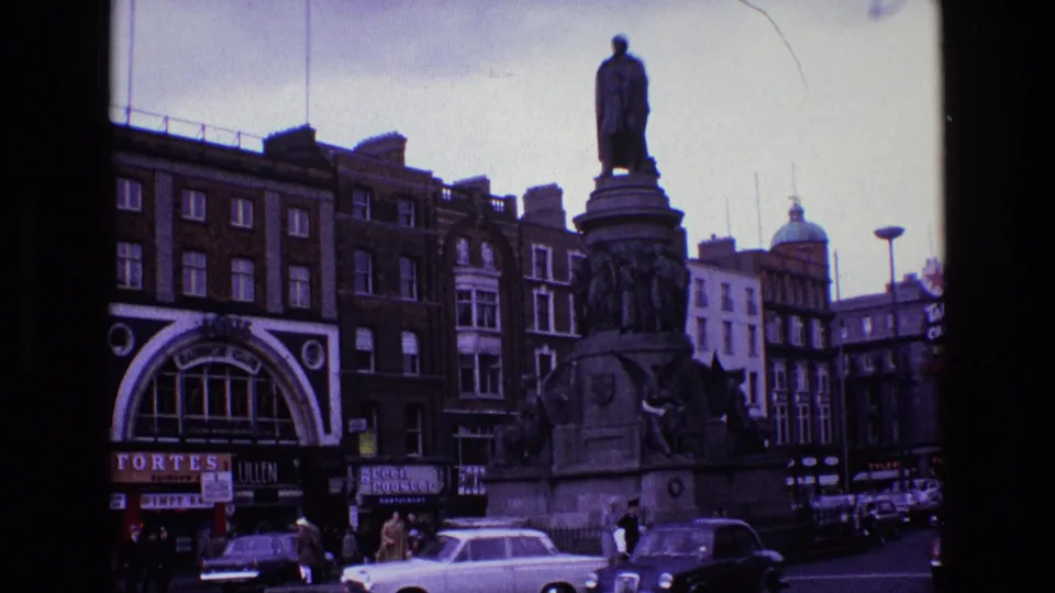 1969 Ireland city square view with a large statue, pedestrians, cars, shopfronts, and surrounding buildings seen through a moving vehicle.