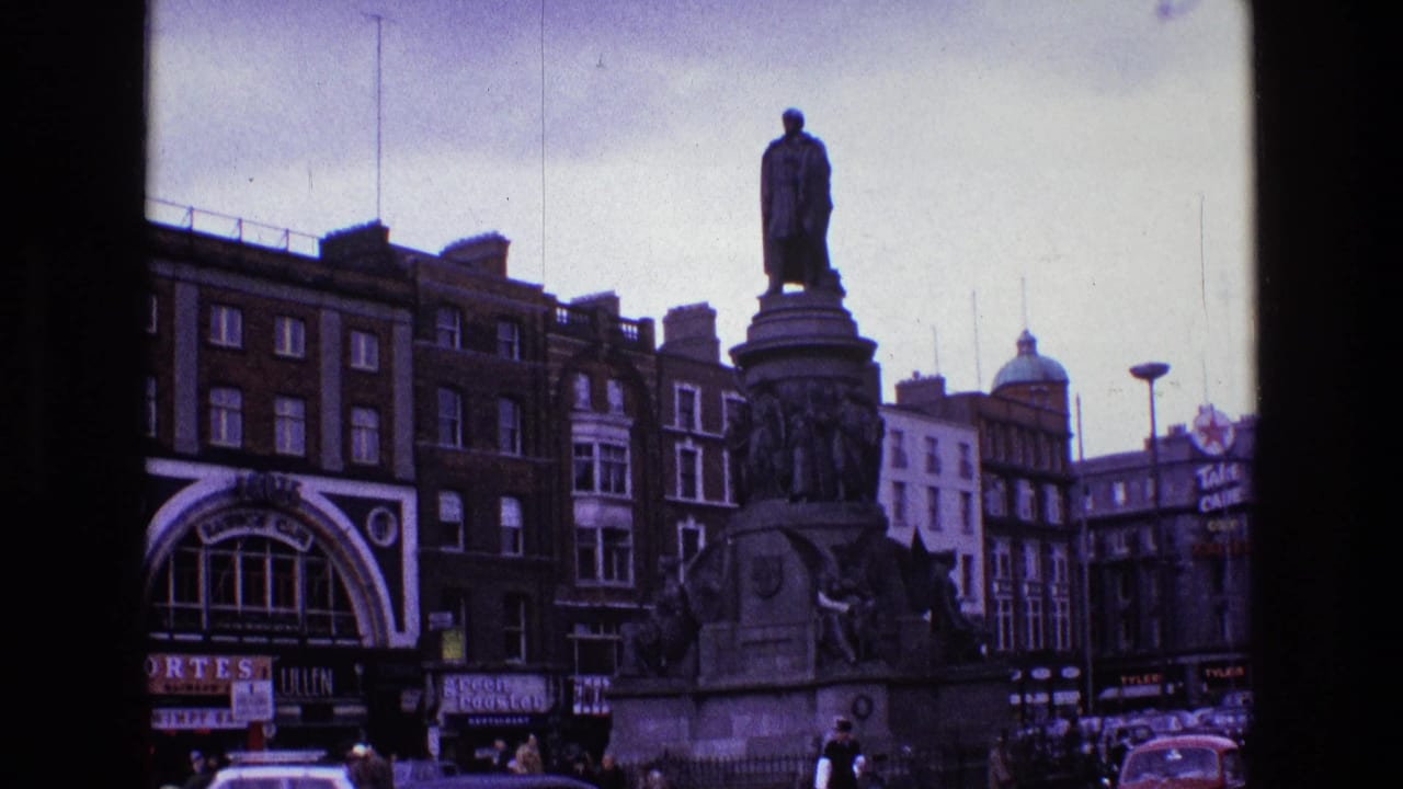 City monument with a standing statue, surrounding buildings, shop signs, cars, and pedestrians.