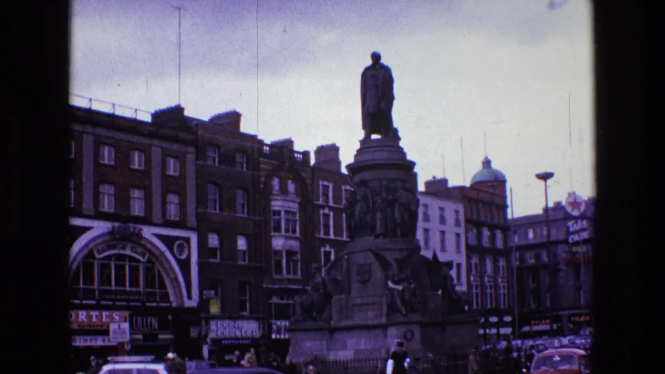 1969 Ireland home-movie frame of a large city monument in a square, with brick buildings, shopfront signage, classic cars, and pedestrians.