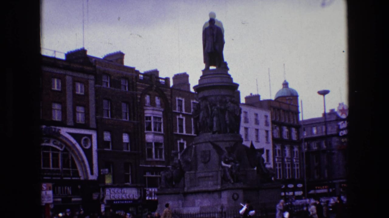 Wide view of a monument in a city square with buildings, cars, pedestrians, and visible signage.