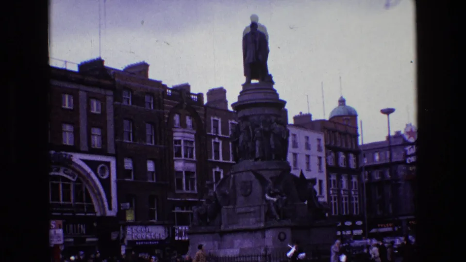 1969 Ireland street scene showing a tall monument with sculpted figures, nearby storefronts, moving cars, pedestrians, and period urban signage.