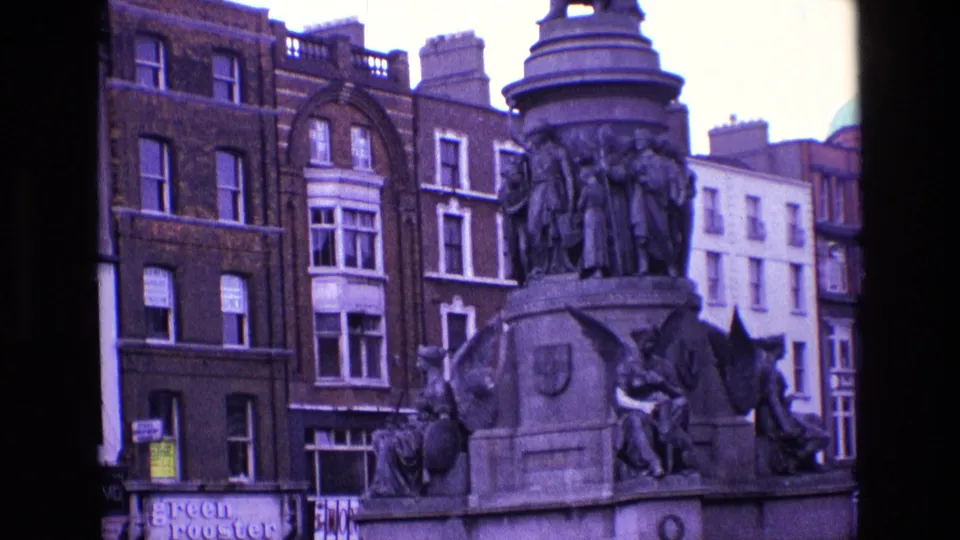 Closer 1969 Ireland home-movie view of the monument's sculpted base, framed against brick buildings, upper windows, and partially readable shopfront signs.