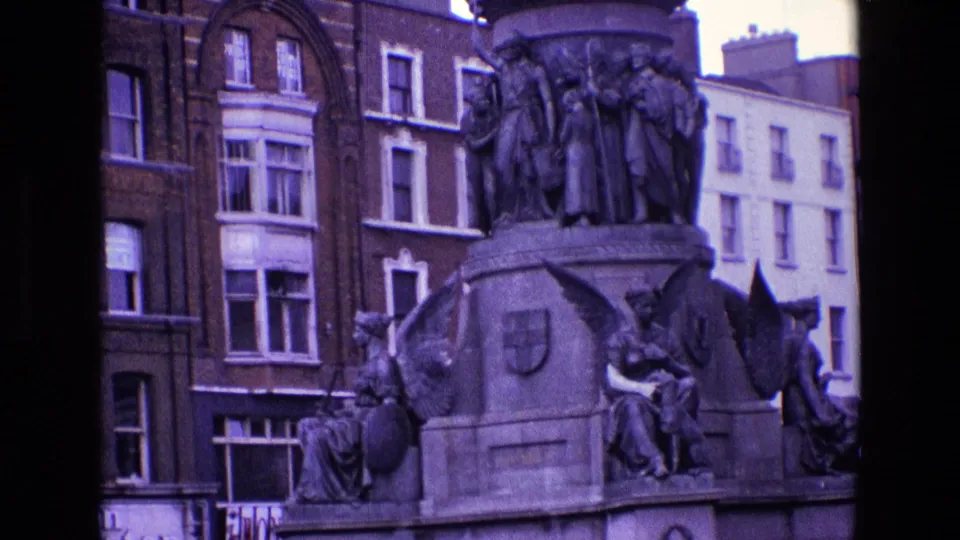 1969 Ireland frame focused on the monument's carved figures and shield details, with brick facades, windows, and street-level signs behind.