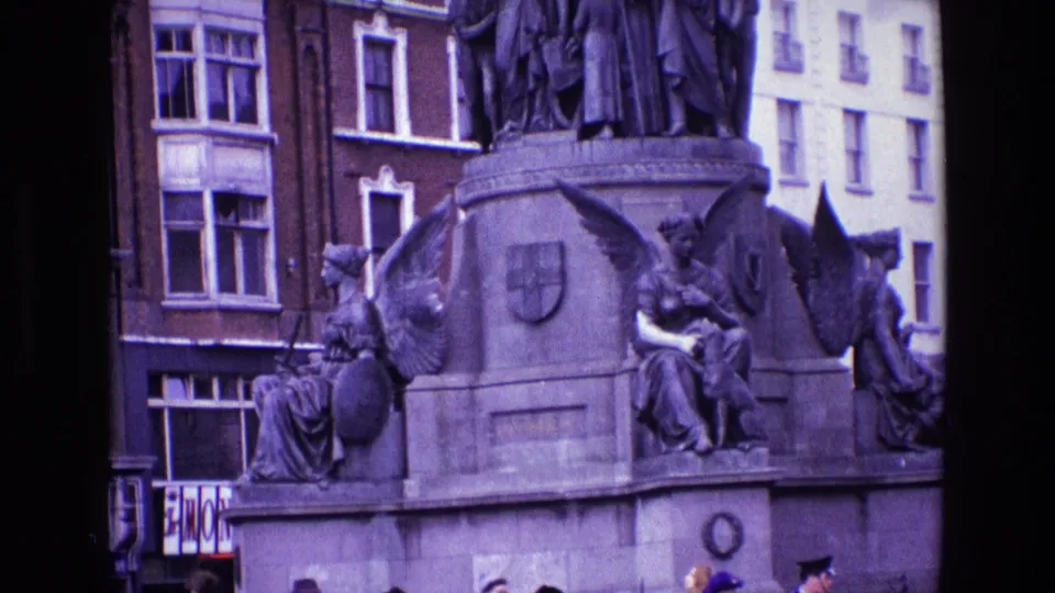1969 Ireland 8mm frame showing a stone civic monument with winged figures, nearby buildings, and partial pedestrians below.