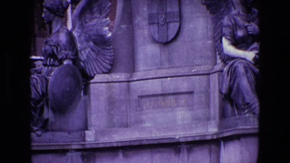 Close 8mm view of the Irish monument base, showing winged sculpture, a shield emblem, and worn engraved stonework.