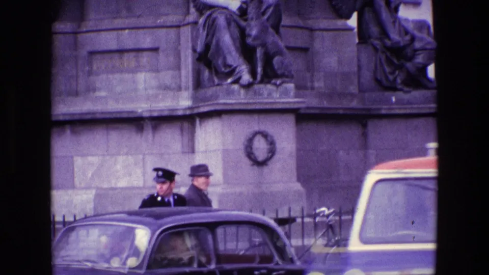 1969 Ireland street frame showing the monument base with a police officer, a man in a hat, vintage cars, and railings below.