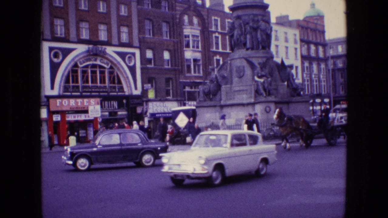 Classic cars move through a 1969 Irish city intersection with a horse carriage, pedestrians, storefronts, and a stone monument behind them.