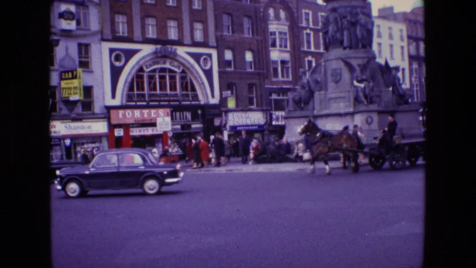 A black sedan passes through a 1969 Ireland street scene with pedestrians, shop signs, a horse carriage, and a sculpted stone monument.