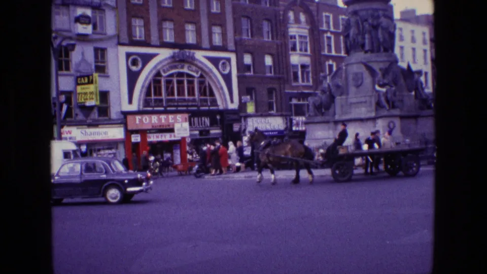 In 1969 Ireland, a horse-drawn cart moves through a city intersection with classic cars, pedestrians, storefronts, and a large stone monument.