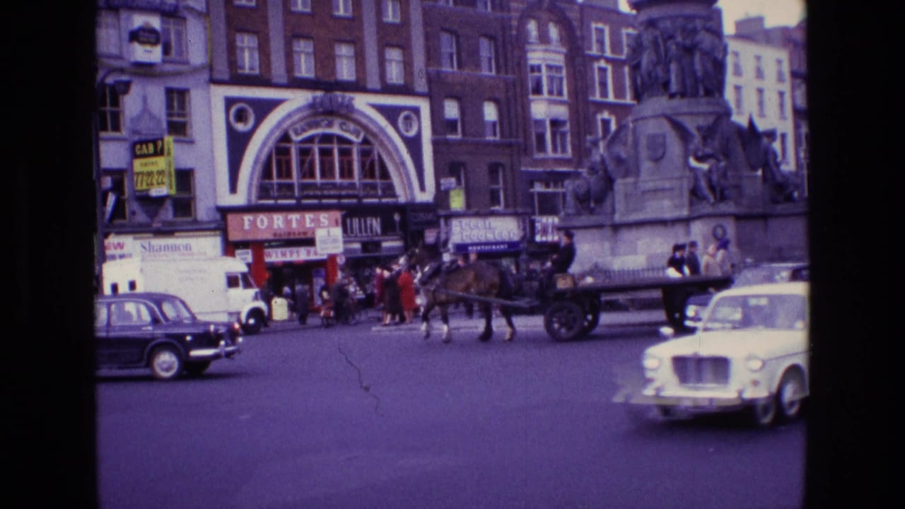 1969 Ireland street scene with classic cars, a horse-drawn cart, pedestrians, storefront signs and a large stone monument.