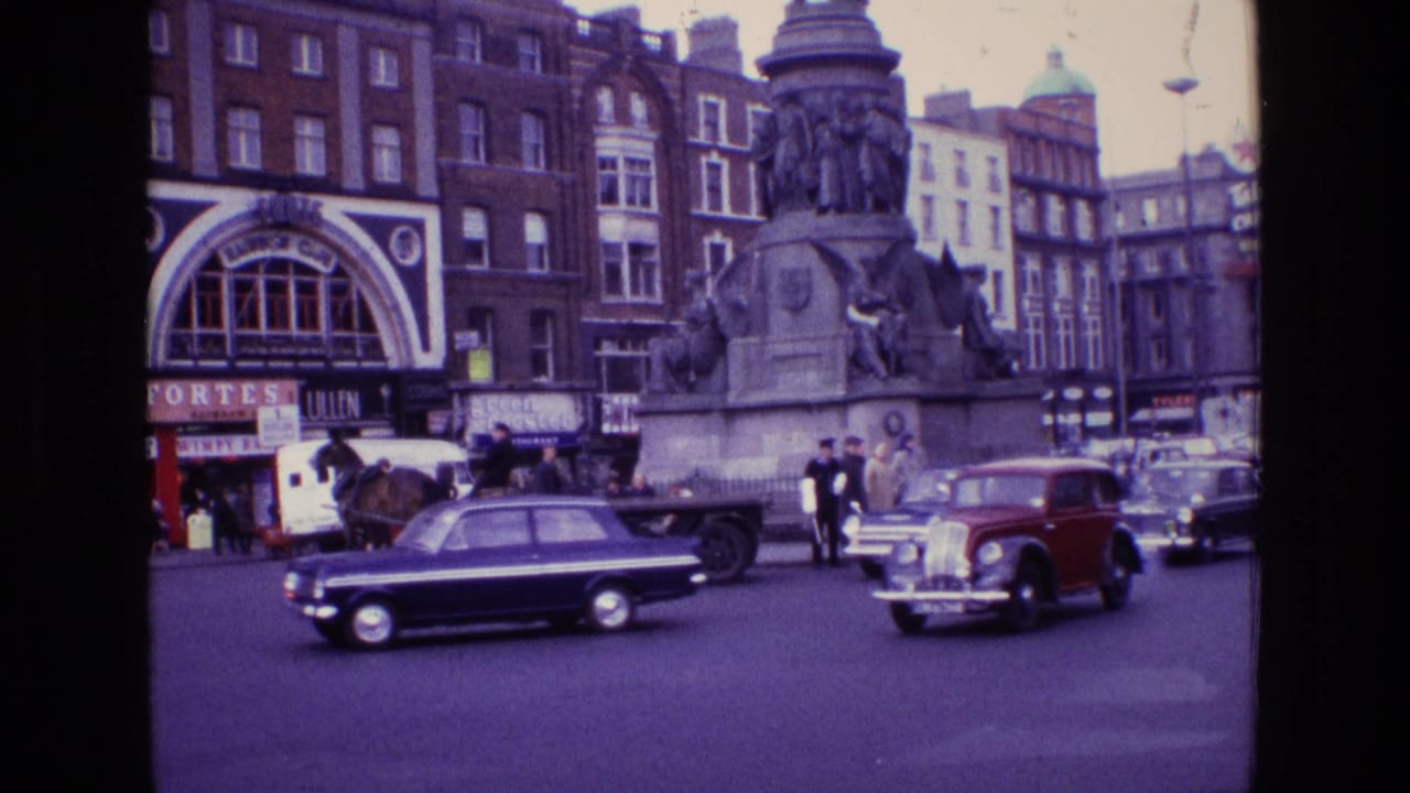 Classic cars pass in front of a stone monument and busy storefronts.
