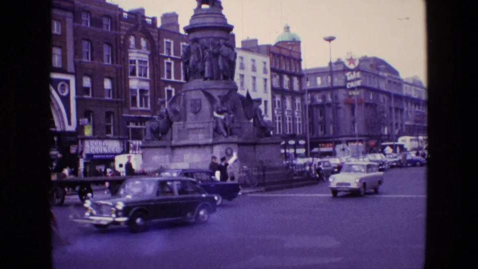 1969 Ireland street footage showing a dark vintage car, pedestrians, storefronts and a large public monument in a city square area.
