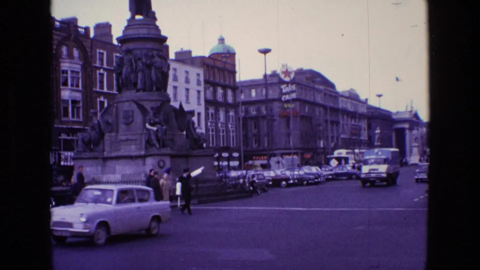 1969 Ireland city view with small cars, pedestrians, a large stone monument, street lamps and rows of commercial buildings.