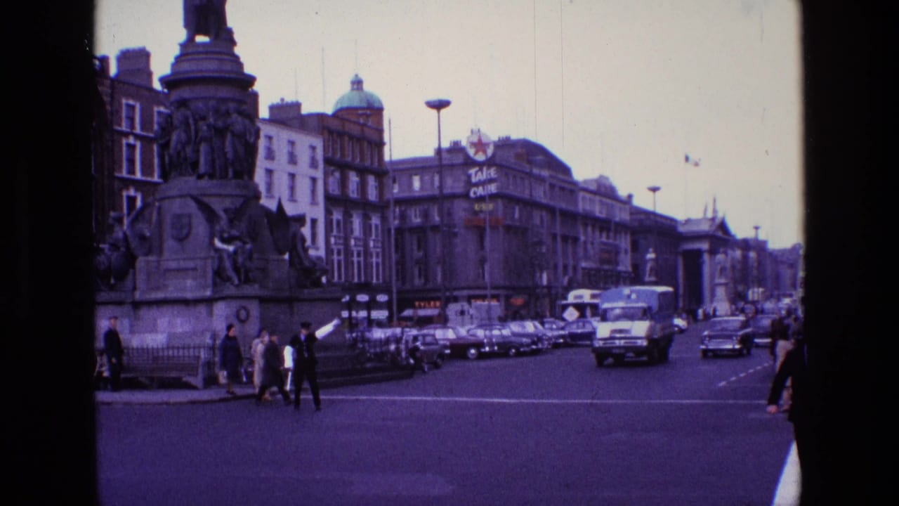 1969 Ireland street view with a stone monument, pedestrians crossing, classic cars, a delivery truck, and commercial buildings.