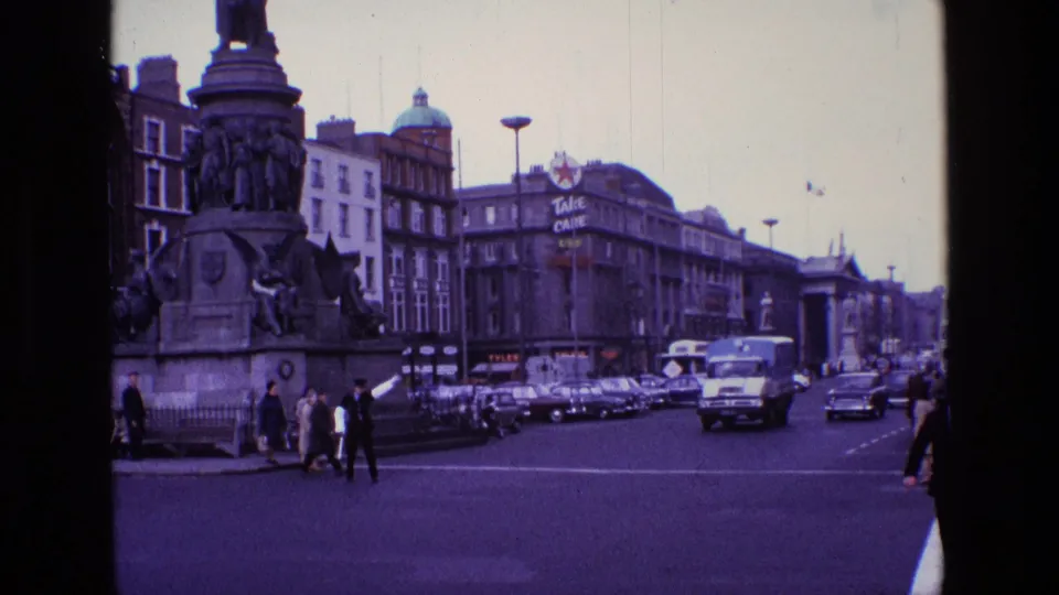 1969 Ireland street view with a stone monument, pedestrians crossing, classic cars, a delivery truck, and commercial buildings.