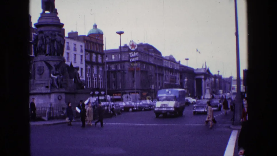 1969 Ireland intersection with pedestrians near a monument, a white delivery truck, classic cars, and shopfront buildings.