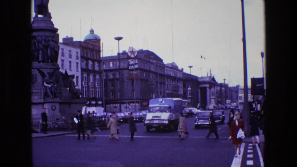 1969 Ireland street frame with a woman in a brown coat crossing near classic cars, a delivery truck, pedestrians, and a monument.