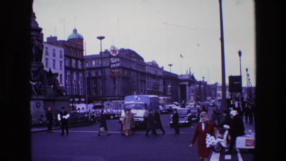 1969 Ireland city street with pedestrians crossing, classic cars, a white delivery truck, a stone monument, and a visible commercial sign.