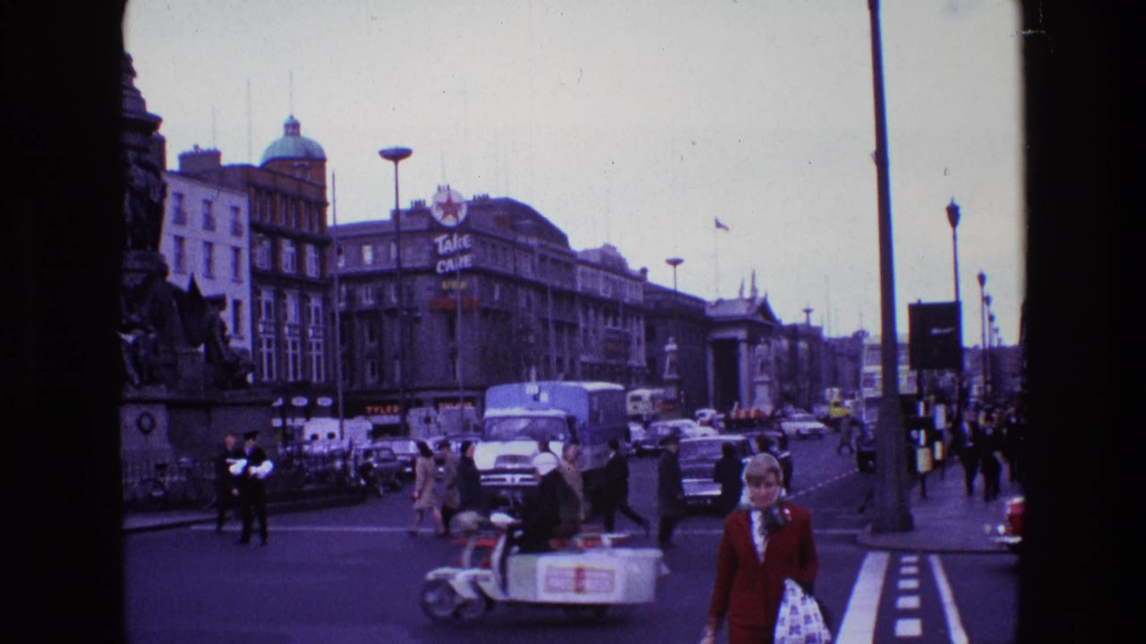 1969 Ireland home-movie view of pedestrians crossing among cars and vans near a large city monument and commercial signage.